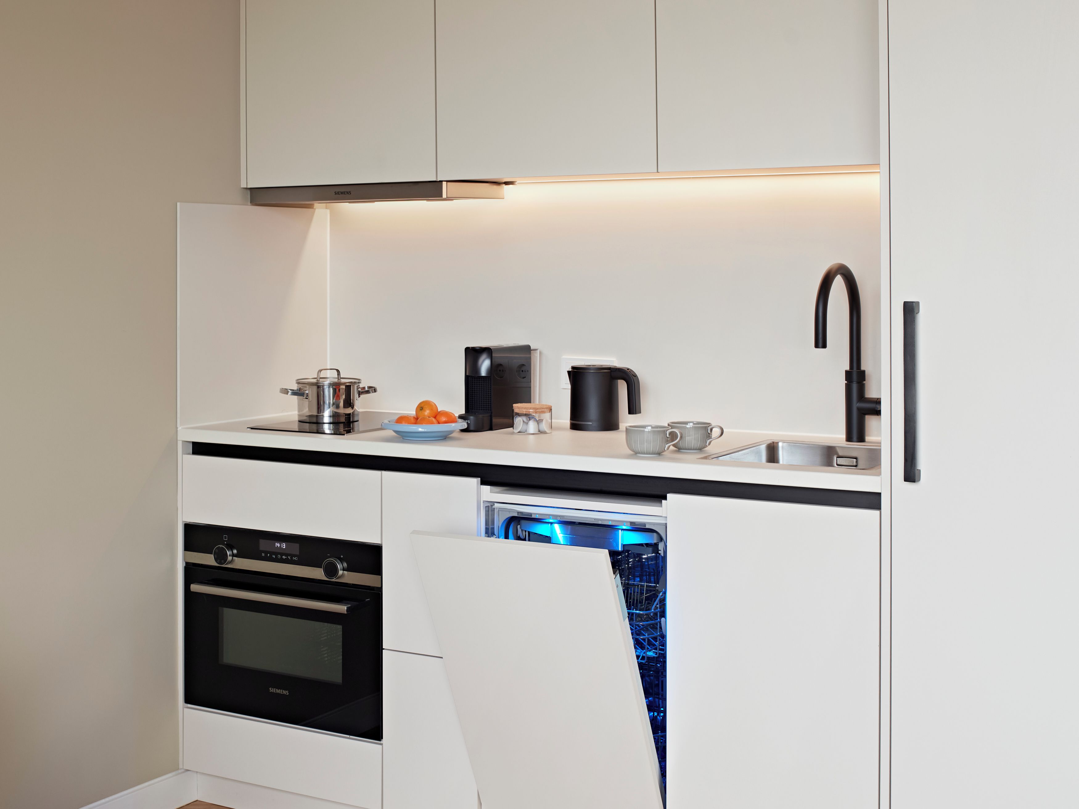 A compact kitchen with white cabinetry, a built-in oven, a dishwasher, and a coffee maker.