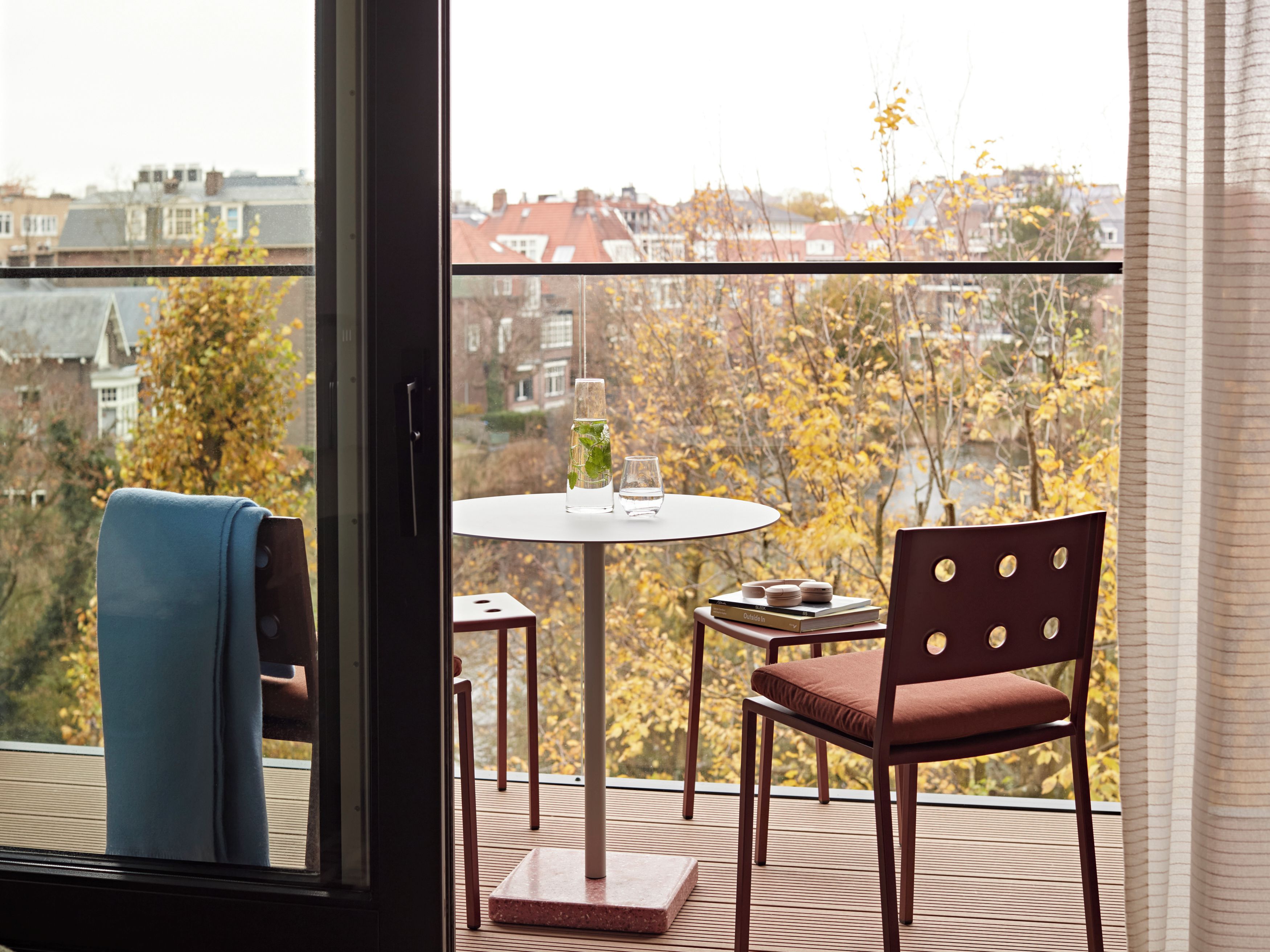 A balcony with a table and chairs, a view of autumn trees, and a building in the background.