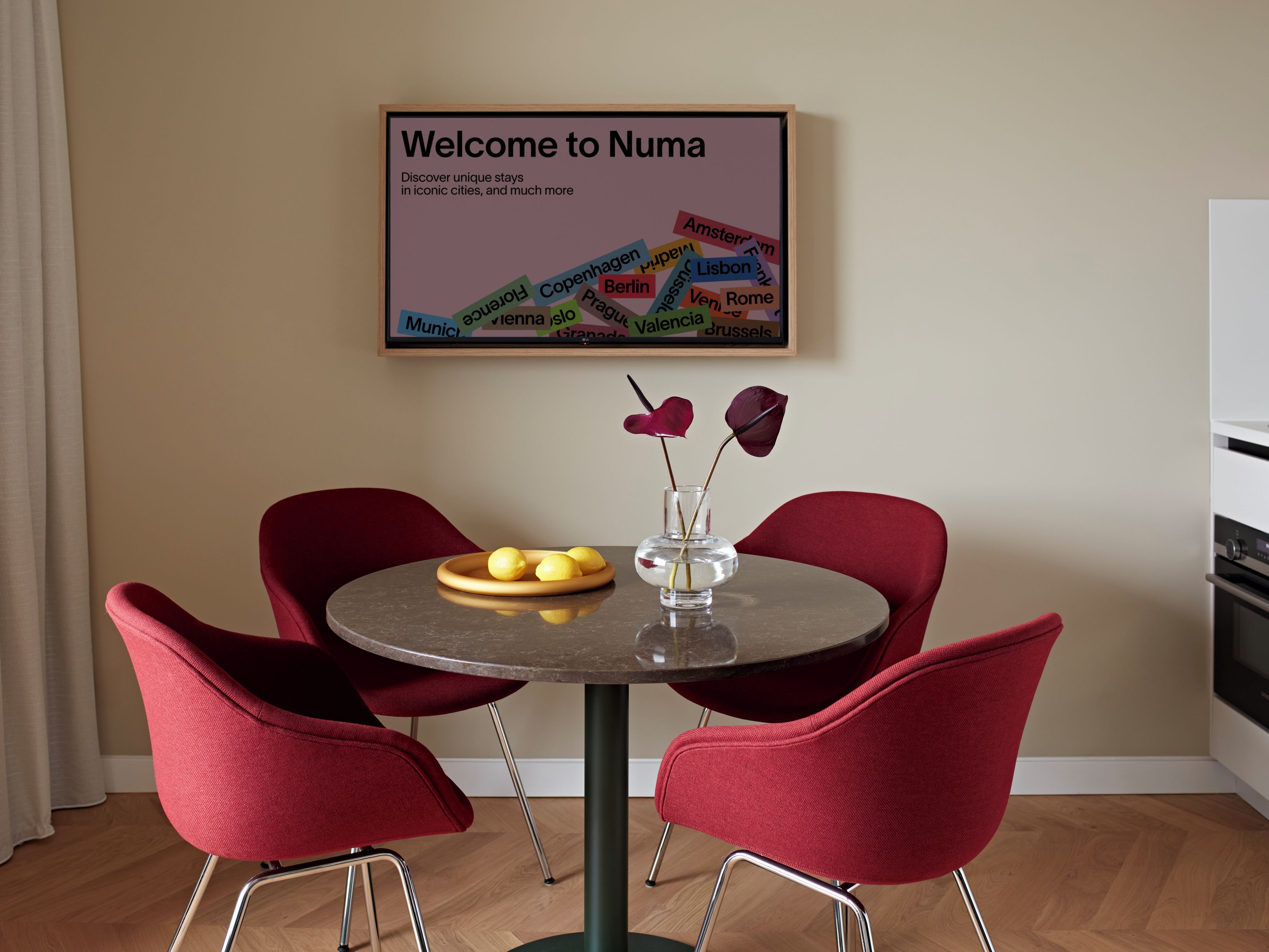 Een eethoek met een ronde tafel, vier rode stoelen, een vaas met bloemen en een lijst met een welkomstboodschap die verschillende steden opsomt.
