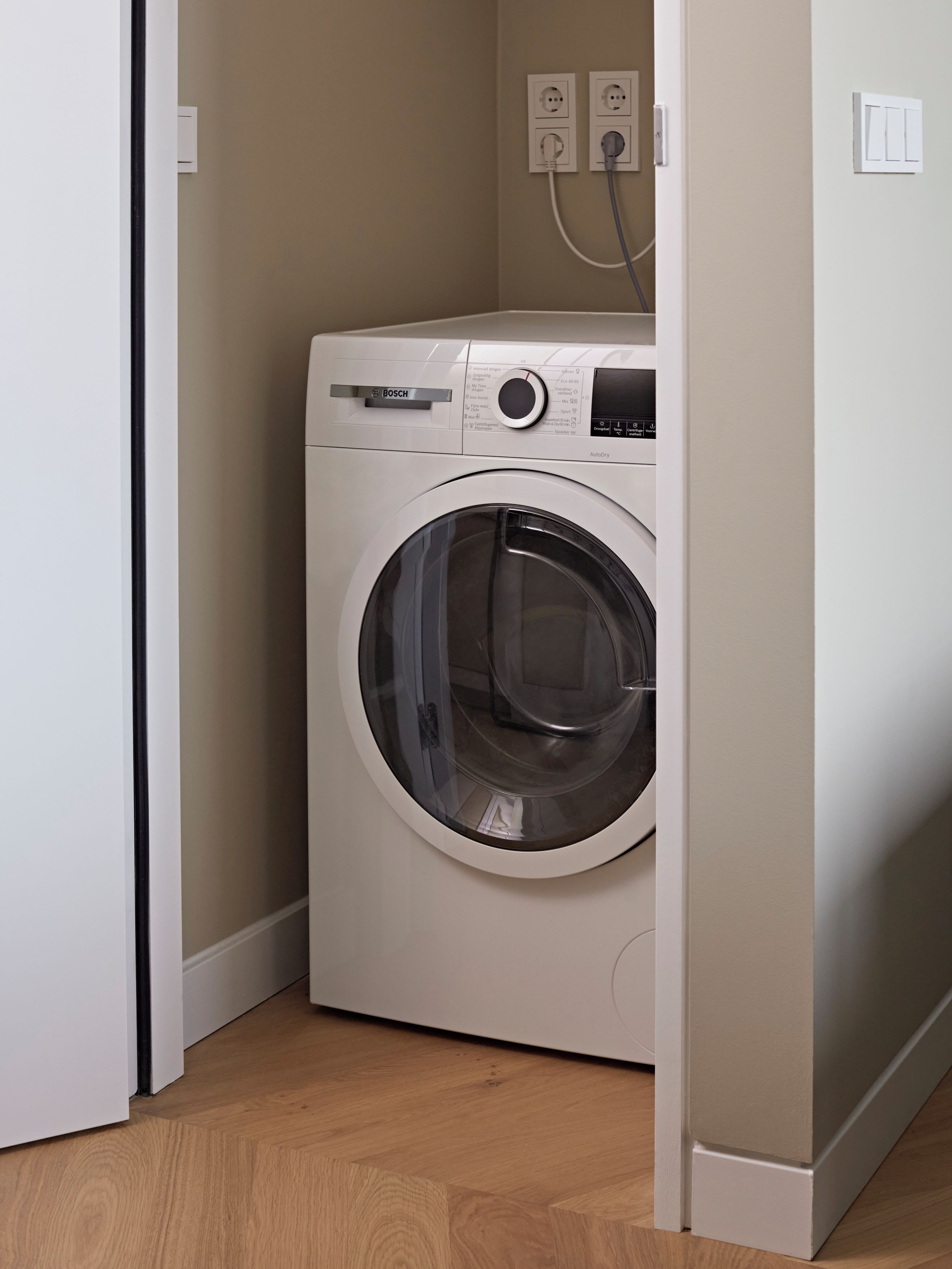 A compact, white Bosch washing machine in a corner of a room with wooden flooring and beige walls.