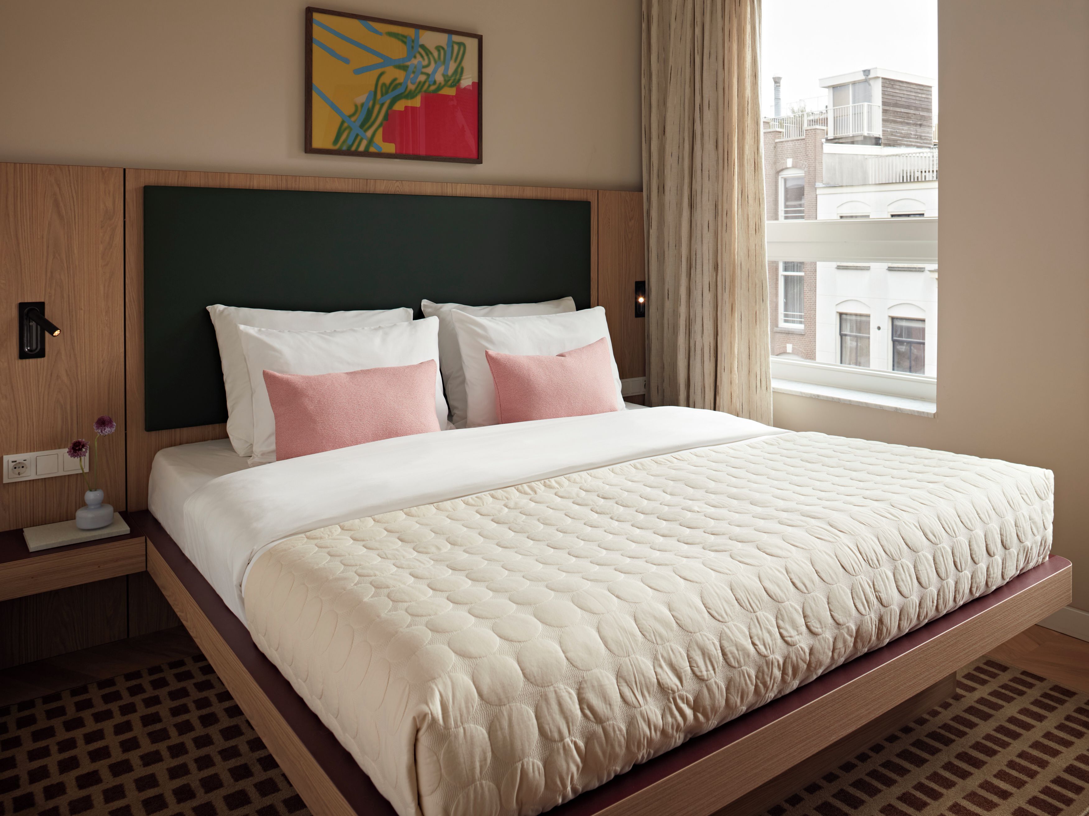 A hotel room with a large bed, a headboard, a nightstand, a vase with flowers, and a window with a view of buildings.