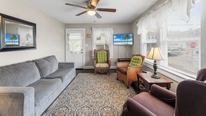 A living room with a gray sofa, wicker chairs, a patterned rug, a ceiling fan, and a TV on the wall. - Shannon A - downstairs (Ocean City)