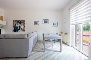 A modern living room with a grey sofa, a dining table with chairs, a radiator, and a large window with blinds. - Apartment 'Rocca Lake House' with Shared Garden, Balcony and Wi-Fi (Angera)