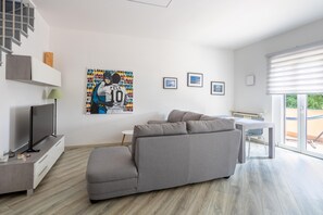 A modern living room with a grey sofa, a flat-screen TV, a small table, and a window with blinds. - Apartment 'Rocca Lake House' with Shared Garden, Balcony and Wi-Fi (Angera)