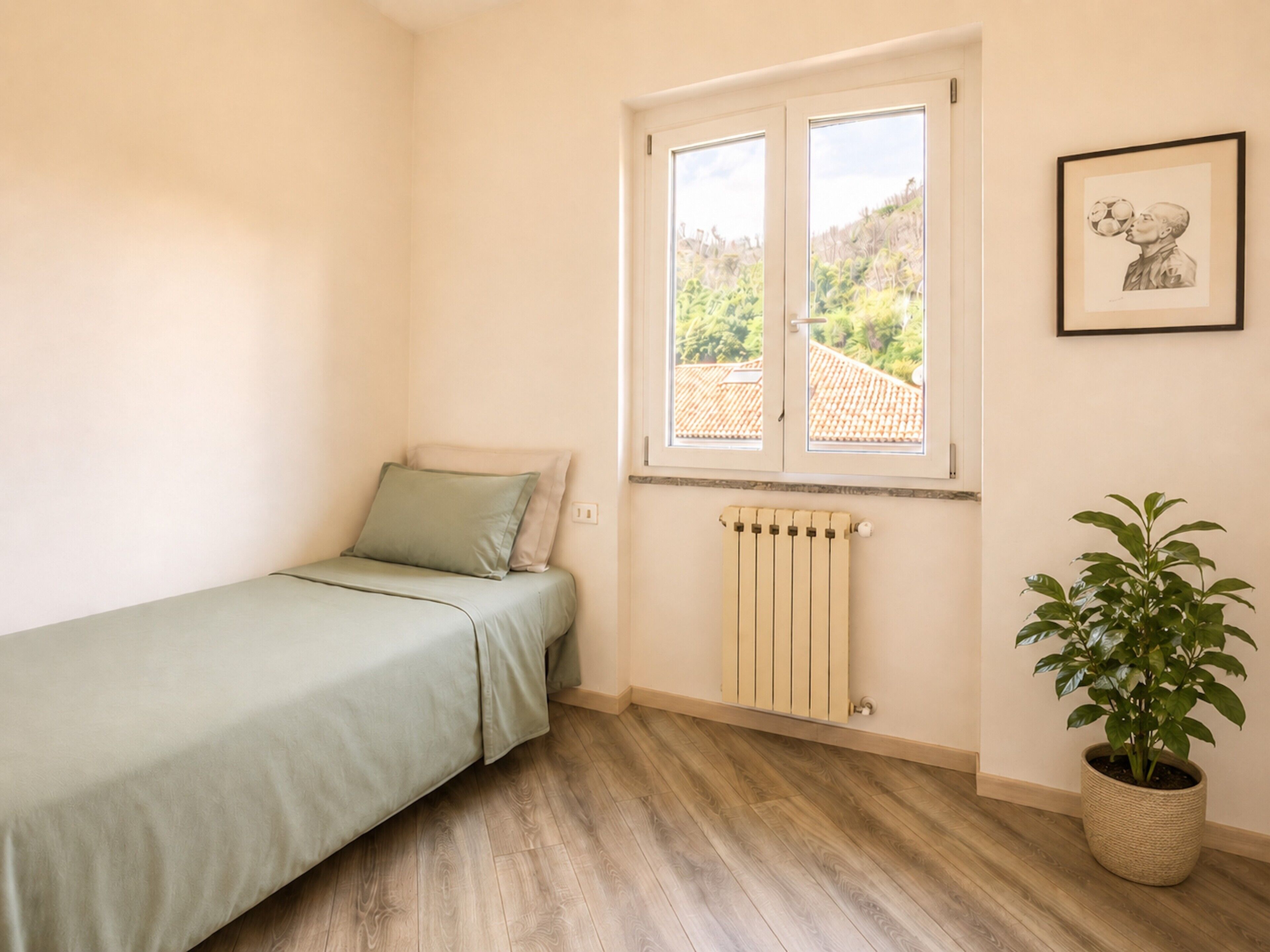 A bedroom with a single bed, a radiator, a window with a view of greenery, and a framed picture on the wall.