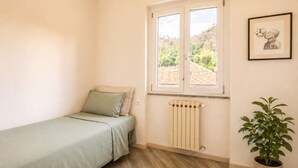 Un dormitorio con una cama individual, un radiador, una ventana con vista a follaje y un cuadro en la pared.