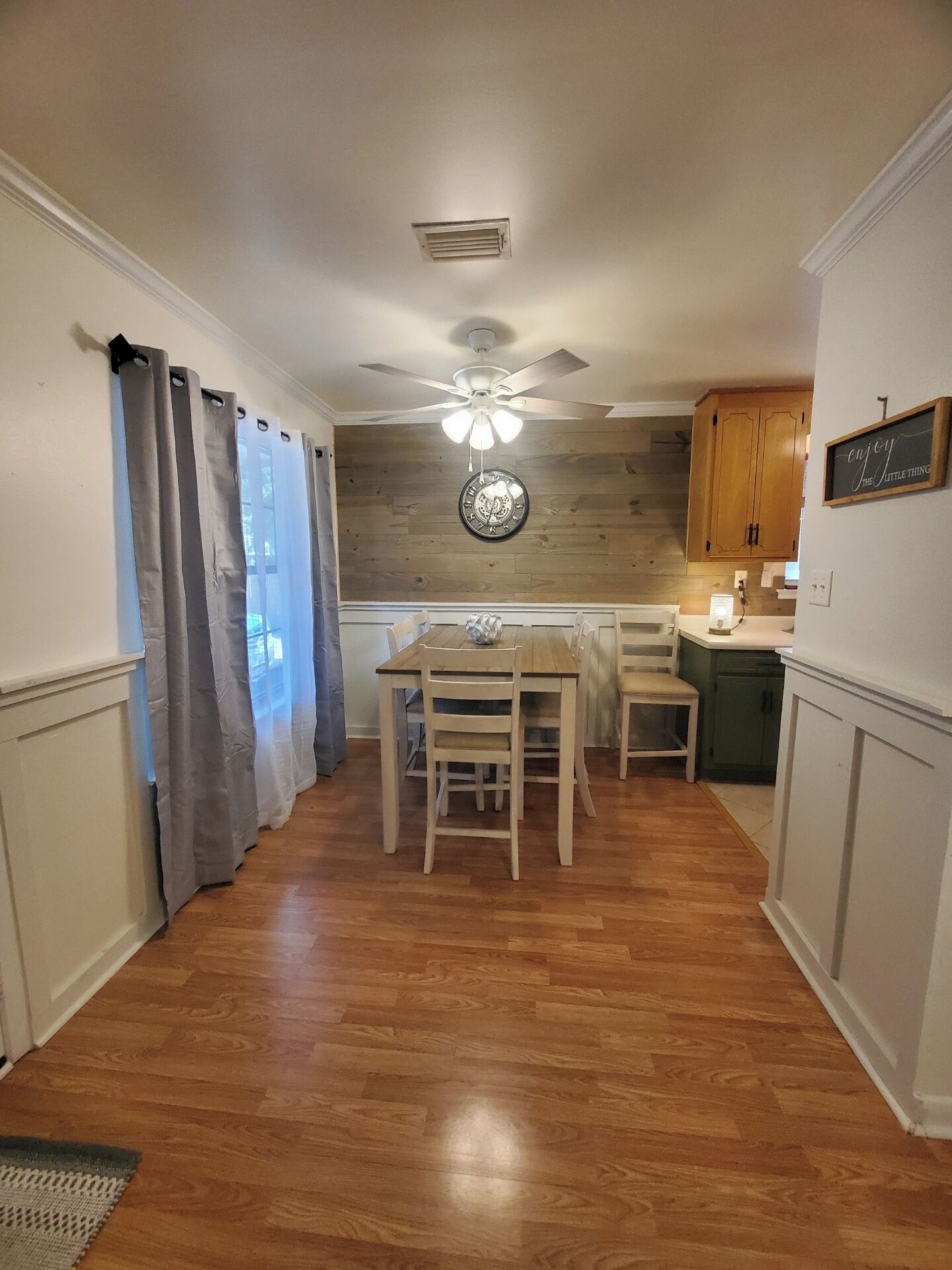 Une cuisine avec des meubles de rangement en bois, une table à manger avec des chaises, un ventilateur de plafond et une horloge au mur.