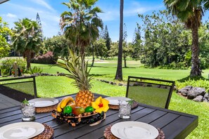A table set outdoors with a tropical fruit arrangement, two chairs, and a golf course in the background. - Keauhou Palena#602 Ground Floor Unit, No Stairs, On Golf Course! (Kailua Kona)