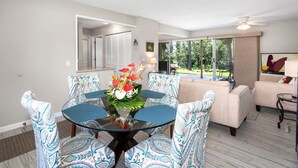 A dining area with a glass table surrounded by patterned chairs, a floral centerpiece, and a view of a garden through large windows.