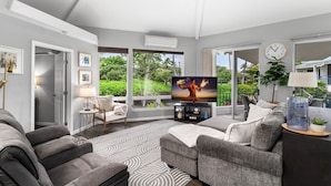 A modern living room with a large TV, comfortable seating, and a view of greenery outside.