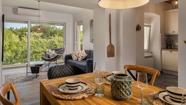 Zona pranzo moderna con tavolo in legno apparecchiato per due persone, una grande lampada a sospensione e vista all'esterno attraverso ampie finestre.