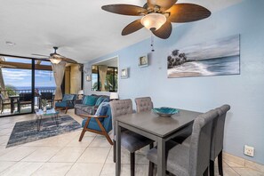 A spacious living area with a dining table, sofas, ceiling fans, and a large window offering a view of the ocean.