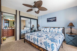 A bedroom with a bed, a ceiling fan, a nightstand, a lamp, a mirror, and a door leading to a bathroom. - Kona Bali Kai 307 Oceanfront– 2BR/2BA Condo with Panoramic Views (Kailua-Kona)