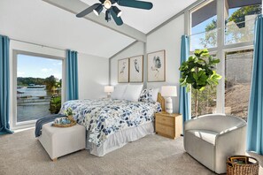 Un dormitorio con un ventanal grande, una cama, una silla, una mesita de noche y un ventilador de techo.