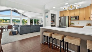 Une cuisine moderne dotée d’un grand îlot central, de meubles en bois et d’appareils électroménagers en acier inoxydable. On y trouve également un coin salon avec un canapé gris et une table à manger.
