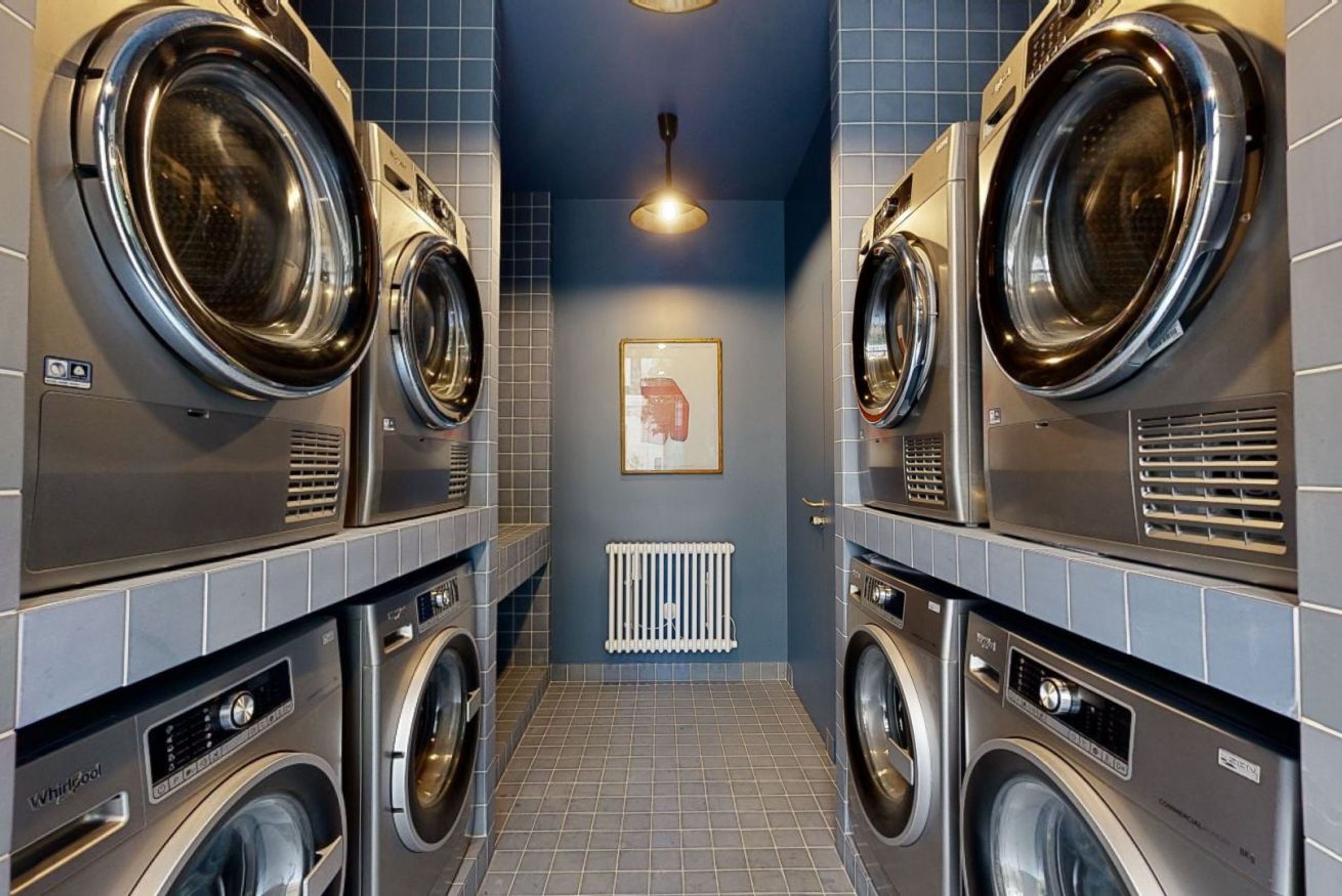 A laundry room with multiple front-loading washing machines and dryers, a tiled floor, and a ceiling light.