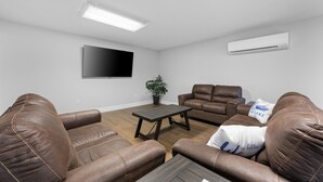 A living room with brown leather sofas, a wooden coffee table, a flat-screen TV mounted on the wall, and an air conditioning unit.