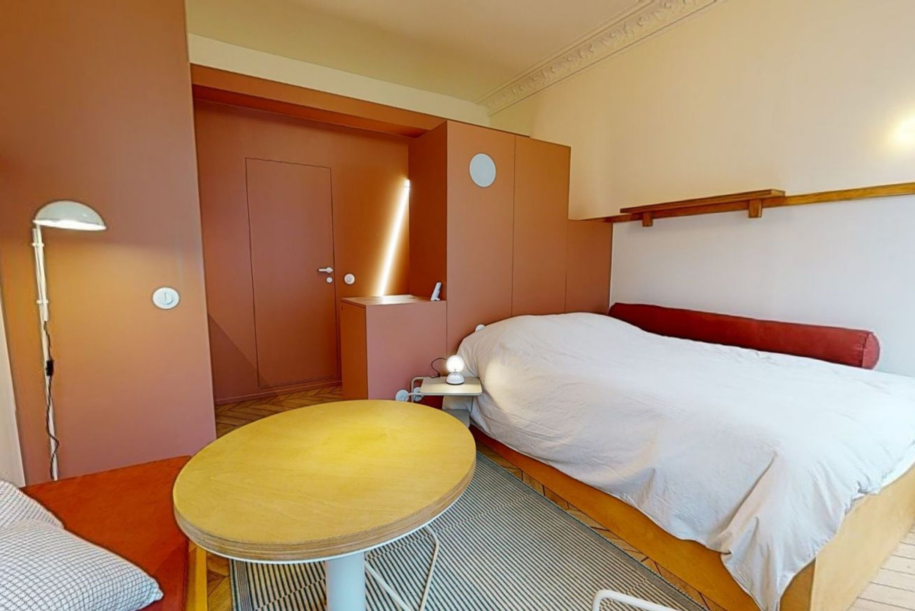 Quarto de hotel compacto com uma cama de solteiro, uma mesinha, um criado-mudo e uma luminária.