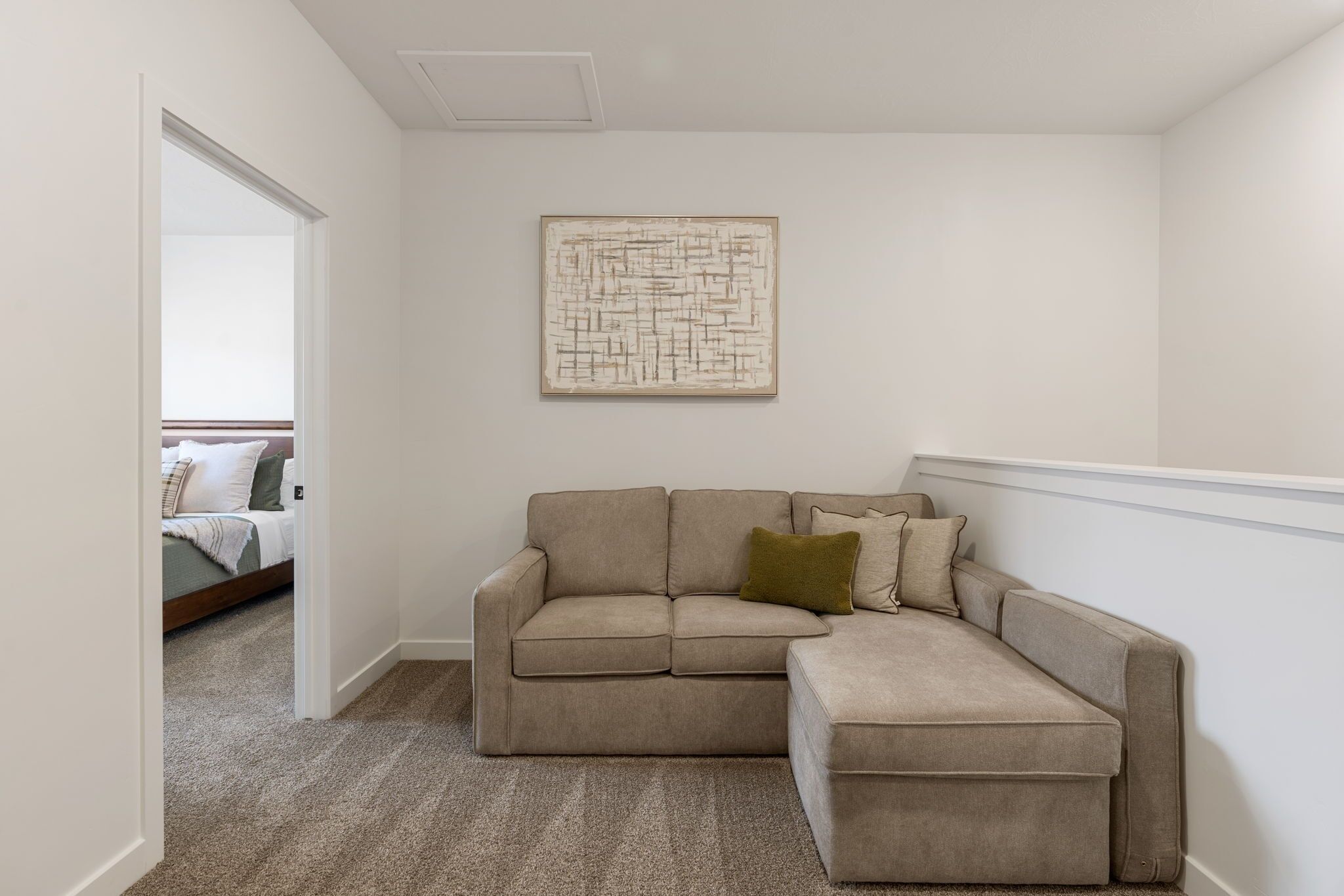 Ein Zimmer mit einer beigefarbenen Eckcouch, einem geometrischen Wandbild und einem Teppichboden.