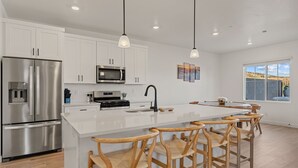 A modern kitchen with white cabinets, stainless steel appliances, a central island with bar stools, pendant lighting, and a view of the outdoors through large windows. - Sunseekers Escape 4 Bedroom Townhouse (Washington)