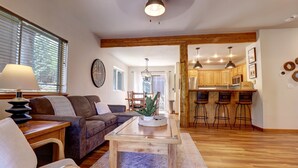 A living room with a sofa, a coffee table, and a lamp. There is a kitchen area with wooden cabinets and bar stools.