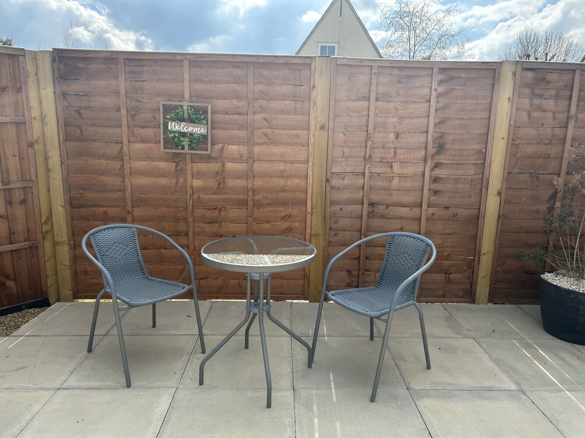 A patio area with two wicker chairs and a small round table, a wooden fence, and a 'Welcome' sign.