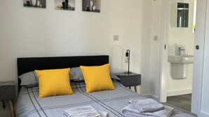 Een net opgemaakt bed met een grijs-zwarte gestreepte dekbedovertrek, twee gele kussens en opgevouwen handdoeken. Boven het bed hangen drie afbeeldingen aan de muur, en er is een badkamer zichtbaar door een geopende deur.