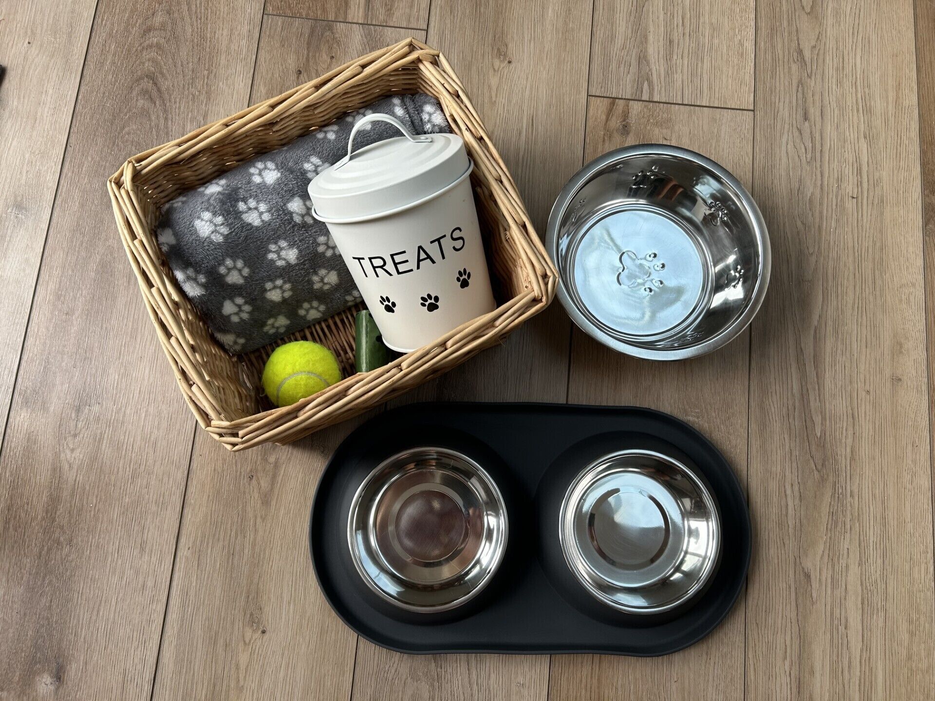 Un panier en osier contenant une tasse blanche sur laquelle est inscrit le mot « GOURMANDISES » et une balle de tennis, ainsi qu’un porte-bougie en métal et deux verres vides sur un plateau noir.