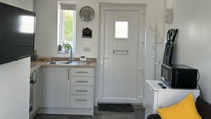 Een compacte keuken met witte kastjes, een spoelbak en een magnetron op een klein tafeltje. Er is een zwarte bank met een geel kussen, een klok aan de muur en een deur met een raam.