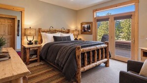 A bedroom with a large bed, wooden headboard, bedside tables, and a view through a glass door. - Alpine 5 Br Retreat / Hot Tub & Mountain Vistas (Big Sky)