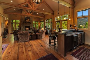 Amplia sala de estar con chimenea, vigas de madera, ventanas grandes y una zona de cocina con taburetes de bar.