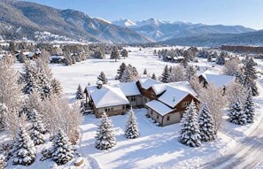 Schneebedeckte Landschaft mit einem großen Gebäude, umgeben von schneebedeckten Bäumen und Bergen im Hintergrund.