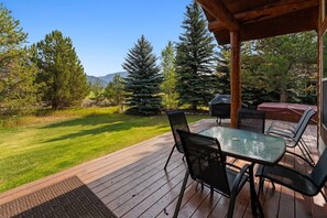 Une terrasse en bois avec une table et des chaises en verre, entourée d’une pelouse bien entretenue et de grands arbres à feuilles persistantes.