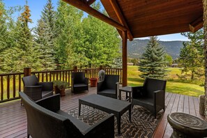 A covered outdoor deck with wicker furniture, a stone planter, and a view of a forested area with mountains in the background.
