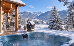 Eine Holz-Blockhütte mit Whirlpool und Sitzbereich im Freien, umgeben von schneebedeckten Bäumen und Bergen.