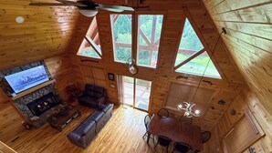 Een gezellig chalet interieur met een stenen openhaard, een leren bank, een eettafel met stoelen en grote ramen met uitzicht op de natuur.