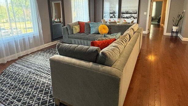 A spacious living room with a large grey sofa, a patterned rug, and a view of a scenic landscape through the windows.