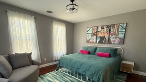 A bedroom with a bed, a grey sofa, a patterned rug, and artwork on the wall.