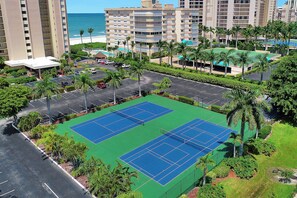 Una cancha de tenis rodeada de estacionamiento y palmeras, con una playa y edificios de gran altura al fondo.