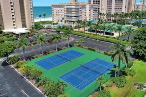 A tennis court surrounded by parking, palm trees, and a beach view.