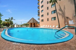 A swimming pool with lounge chairs and a small gazebo, surrounded by a brick walkway and a multi-story building.
