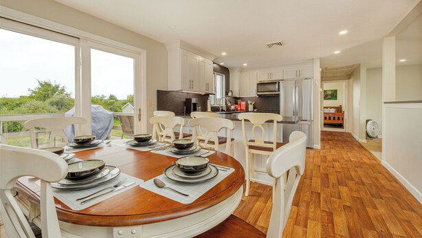 A dining area with a round table set for six, surrounded by white chairs. A kitchen with white cabinets and stainless steel appliances is visible in the background.