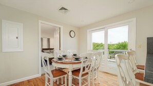 A dining area with a round table and white chairs, a clock on the wall, and a view through a large window.