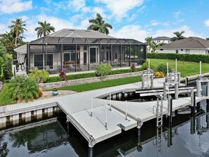 A modern house with a large glass conservatory, a dock extending into a body of water, and a well-maintained garden with palm trees.