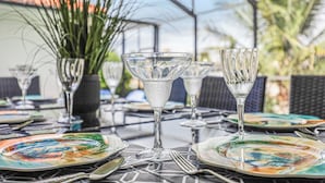 Una mesa dispuesta con cristalería transparente y platos coloridos, rodeada de sillas de mimbre y plantas en macetas.