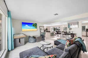A modern living room with a large window, a painting of a beach, and a kitchen in the background.