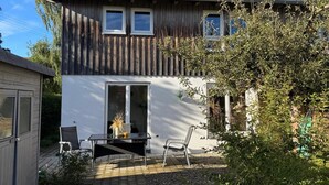 Ein zweistöckiges Haus mit Holzfassade, einer Terrasse mit Tisch und Stühlen und einem Garten.