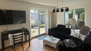 Ein Wohnzimmer mit einer schwarzen Couch, einem weißen Couchtisch, einer schwarzen TV-Konsole und einem großen Fenster mit Blick auf einen Garten.