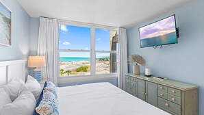 A bedroom with a bed, a dresser, a TV, and a window with a beach view. - Charming Coastal Retreat Mag House 410 | Steps to the Beach + Pool Access! (Destin)