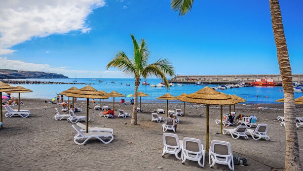 Ein Strand mit Liegestühlen und Sonnenschirmen, einem wolkenlosen blauen Himmel und Booten im Wasser.
